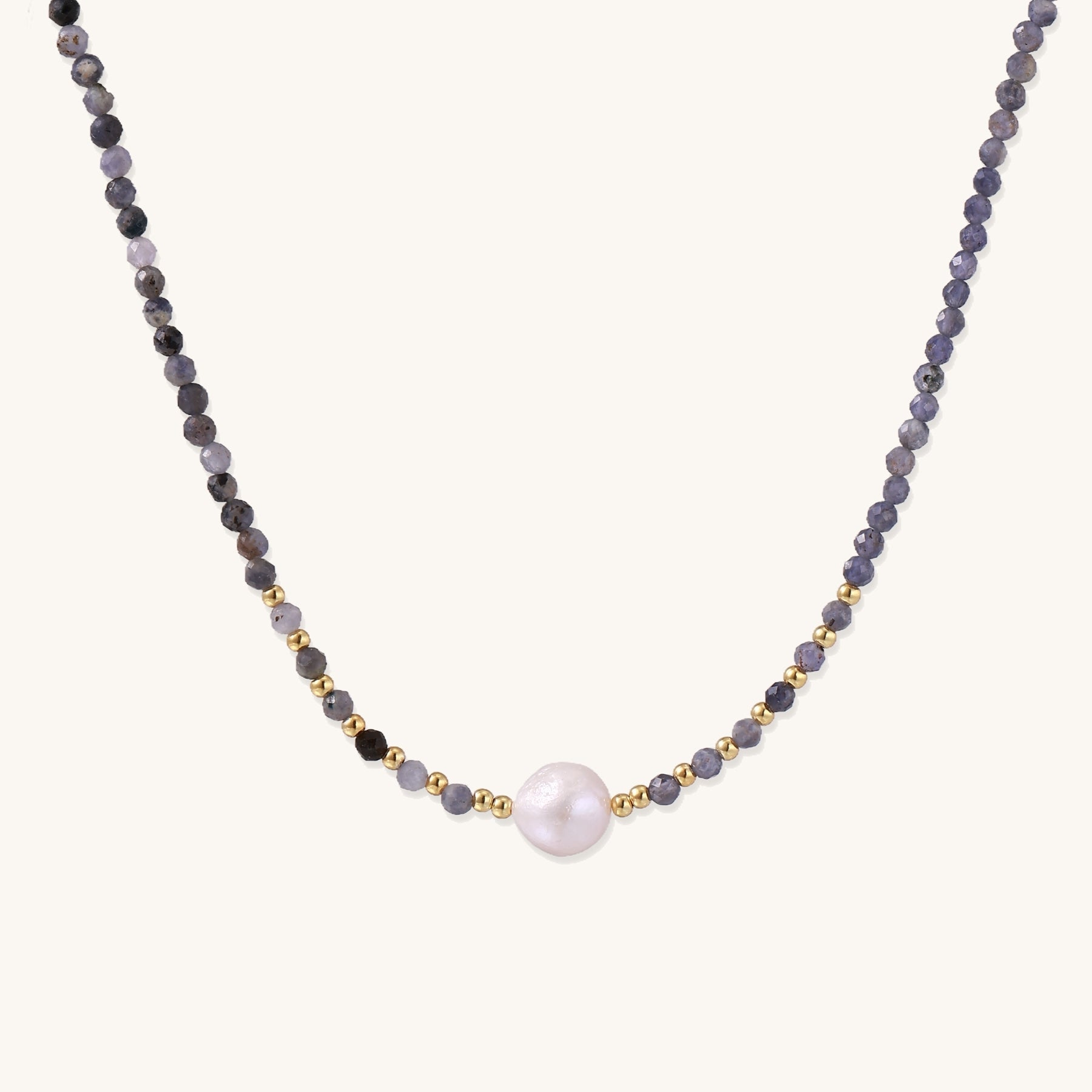 Delicate beaded necklace with violet iolite stones, gold spacers, and central white freshwater pearl on neutral background.