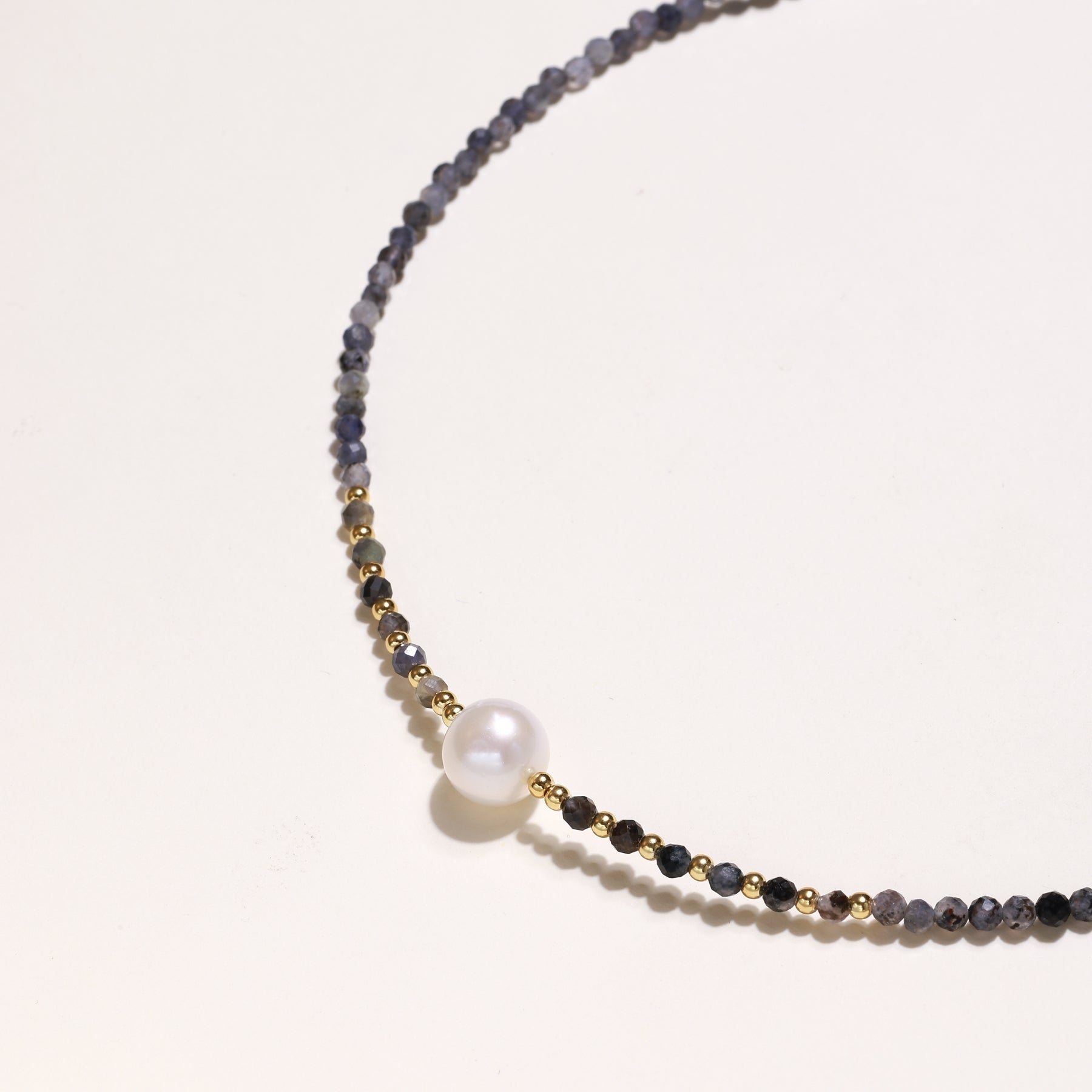 Delicate beaded necklace with violet iolite stones, gold spacers, and central white freshwater pearl on neutral background.