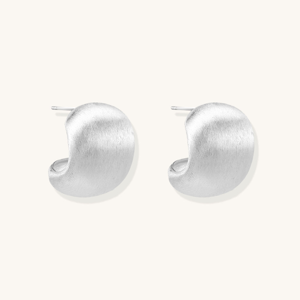 Close-up of chunky sterling silver huggie hoops with brushed matte finish against cream background.