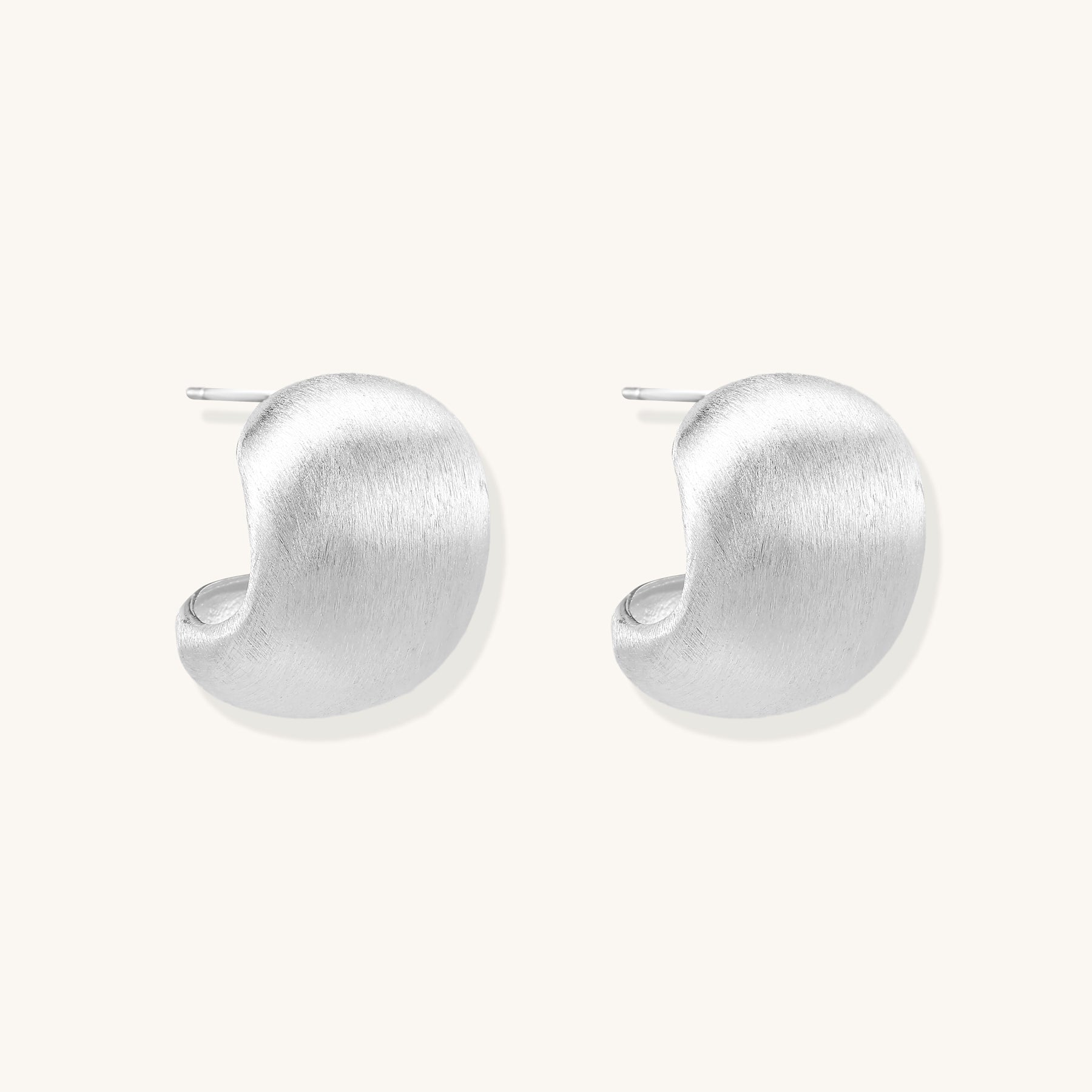 Close-up of chunky sterling silver huggie hoops with brushed matte finish against cream background.