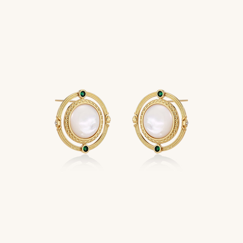 Close-up of gold-plated circular earrings with white mother-of-pearl centers and green zirconia accents on cream background.