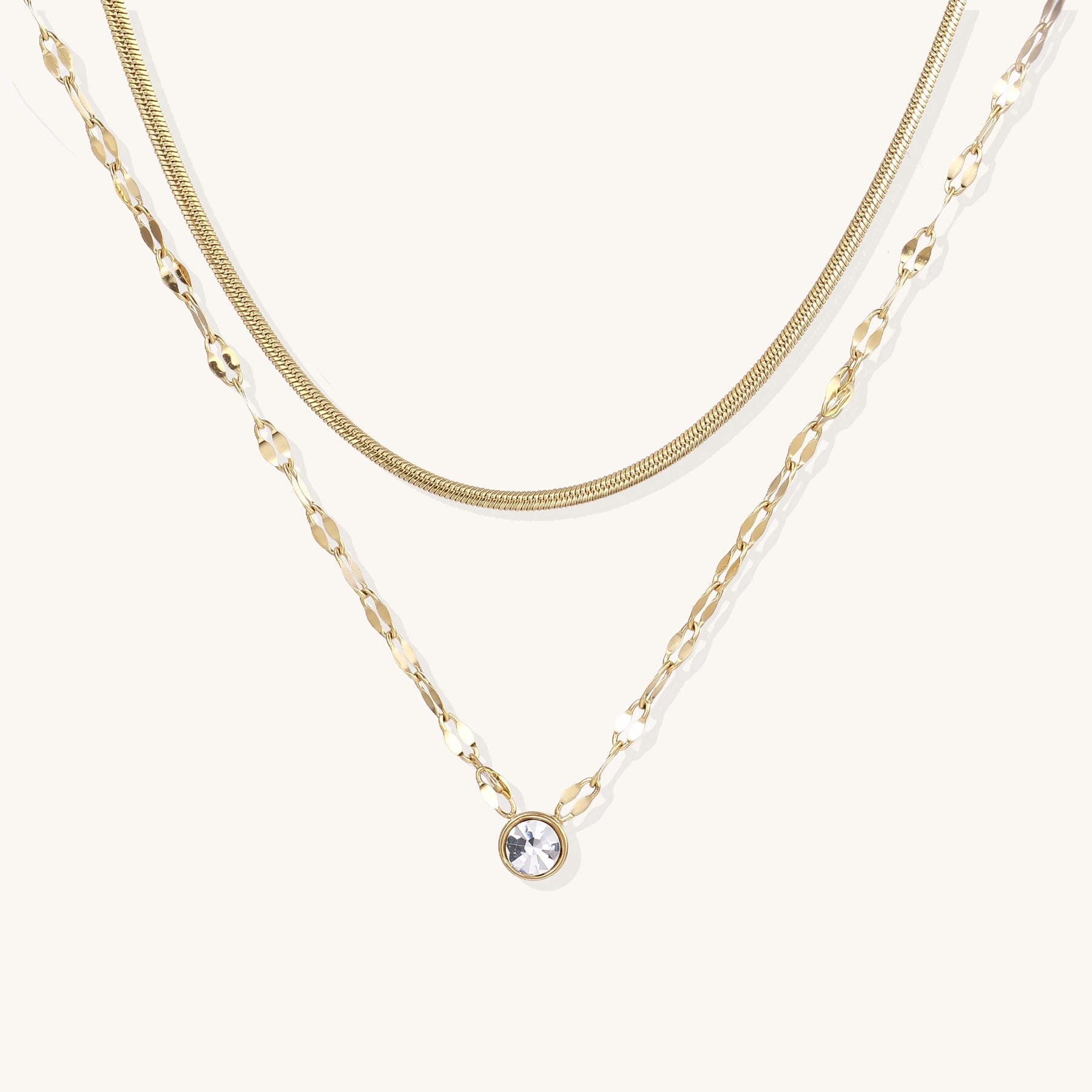 Two layered 14K gold plated necklaces - herringbone chain and delicate chain with circular crystal pendant on beige background.