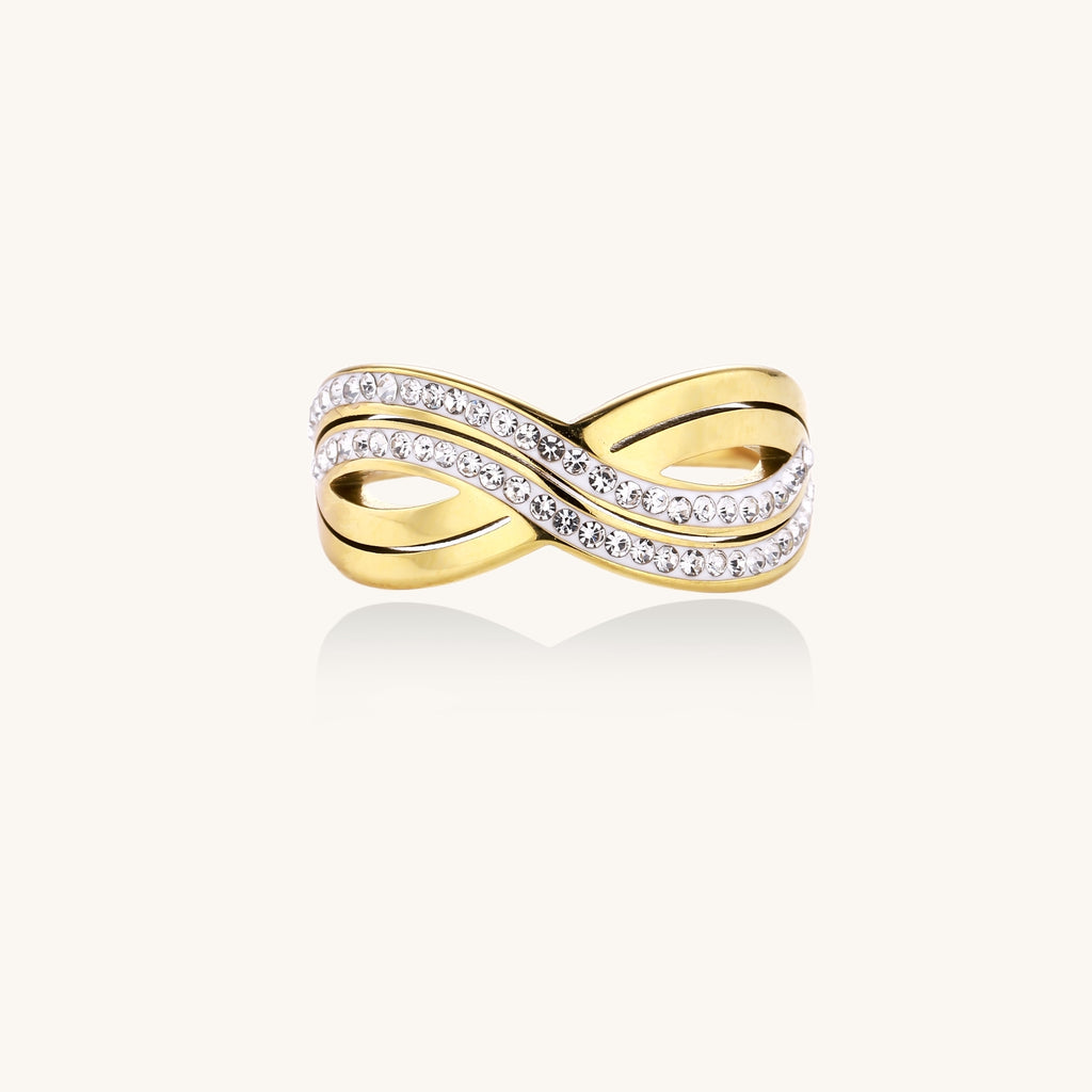 Gold-plated stainless steel ring with crossed infinity bands adorned with sparkling rhinestones against cream background.
