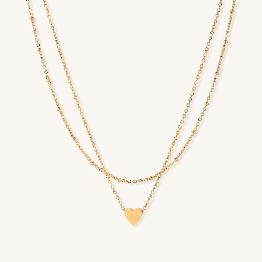 Two-layer gold-plated stainless steel necklace with small heart pendant displayed on beige background