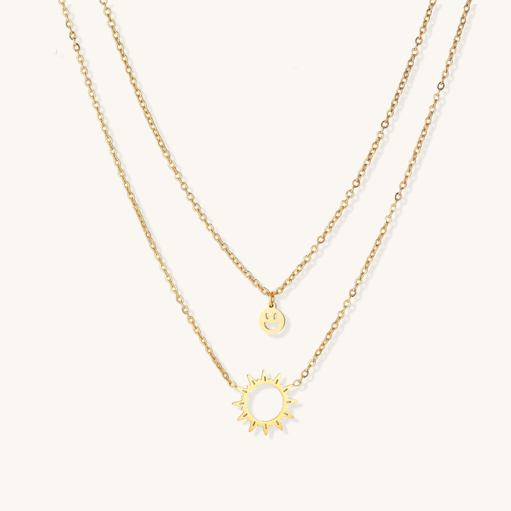 Delicate gold-plated layered necklace with smiley face and sun pendants on white background, showing adjustable chain.